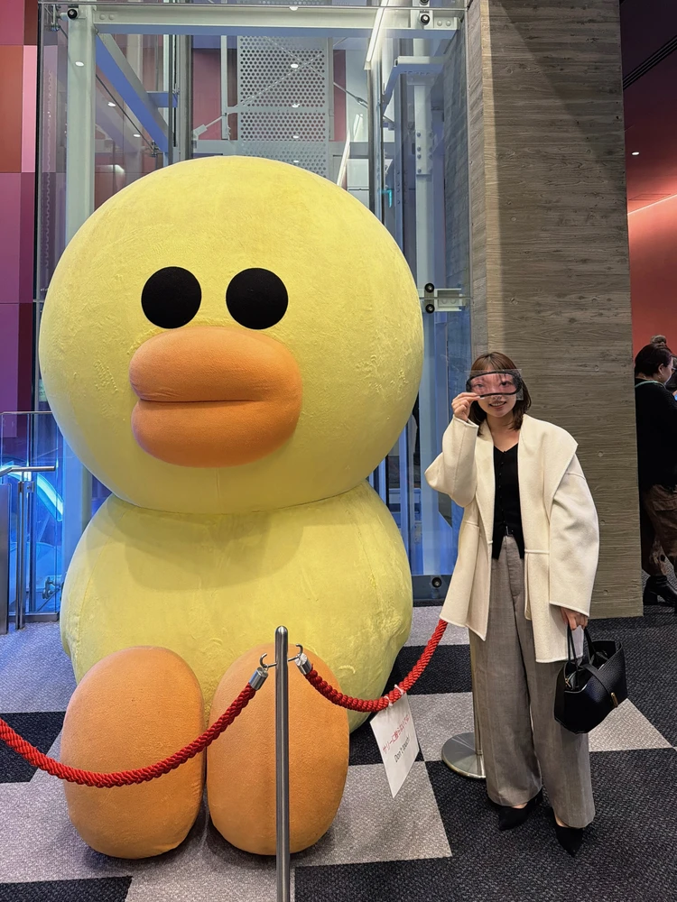 LINEのキャラクター「サリー(黄色い鳥)」の巨大オブジェの隣に立つ様子。手にはS席特典の「永野・くるまの目がプリントされたシュールなアイマスク」を持っている。