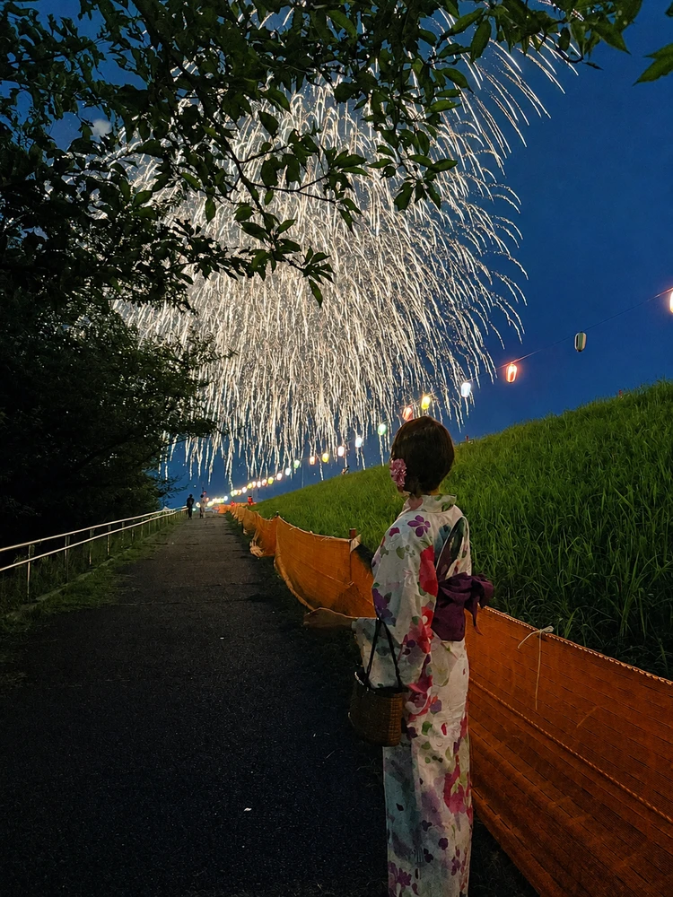 花火が打ち上がる夏の夜、生成り地に色とりどりの花柄が描かれた浴衣を着て歩く女性。深いパープルの帯と低めのまとめ髪で大人っぽく着こなしている