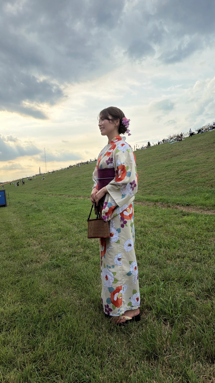 夕暮れの広い芝生の中で、生成り地に赤やオレンジ、紫、ブルーの花柄が描かれた浴衣を着た女性。パープルの帯と花飾り、かごバッグを合わせた大人っぽい浴衣コーディネート